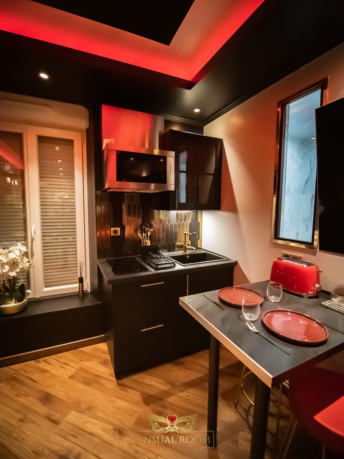 A sleek kitchen is presented with dark cabinets and gold accents. A microwave is mounted above the cooking area, and a red Smeg kettle adds a pop of colour. A reflective mirror enhances depth, while a small table is set with plates and glasses for dining.