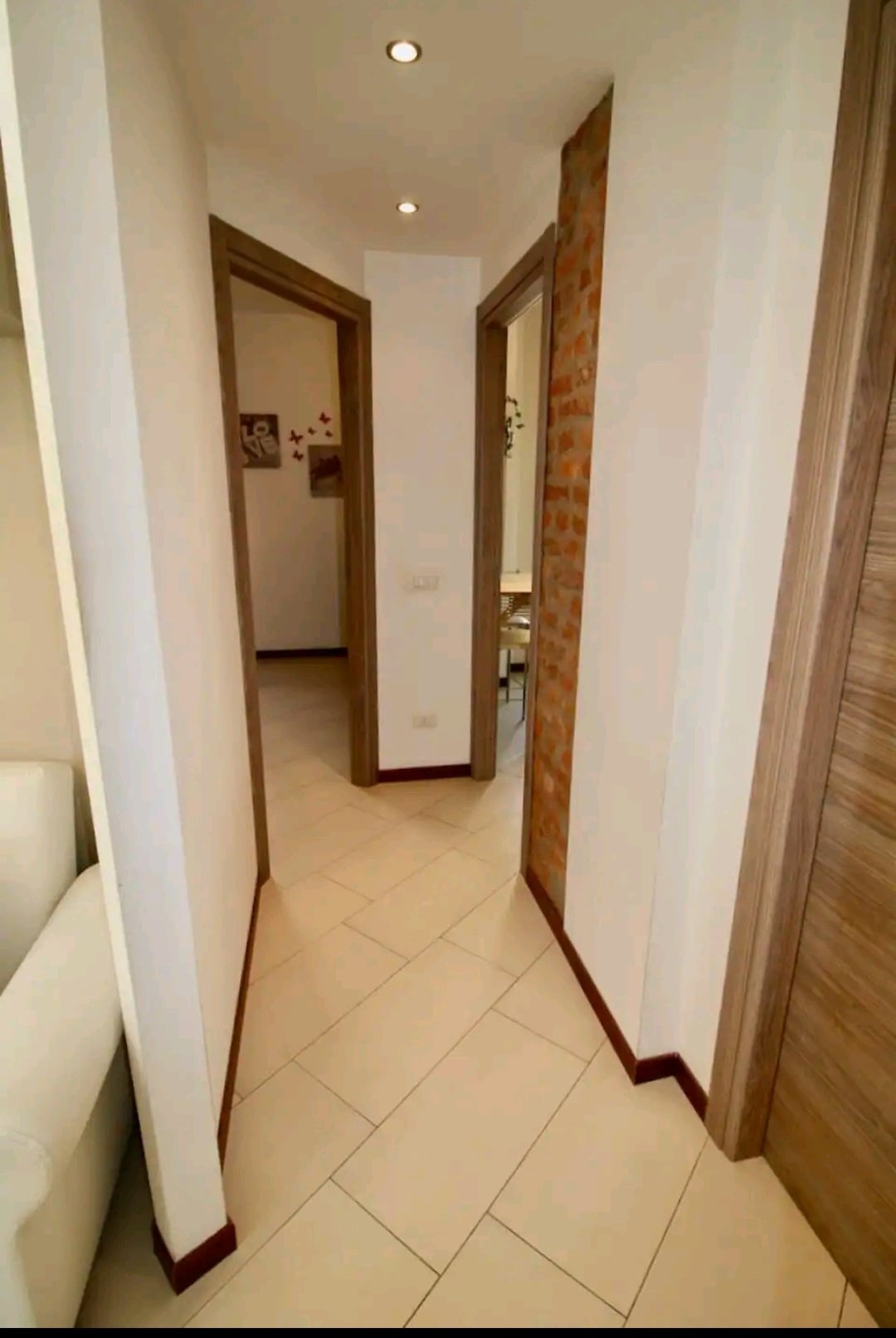 A well-lit hallway features smooth tiled flooring and beige walls. The space is defined by light wooden door frames leading to other areas of the apartment. A hint of exposed brick adds character to the left side, contributing to a welcoming atmosphere.