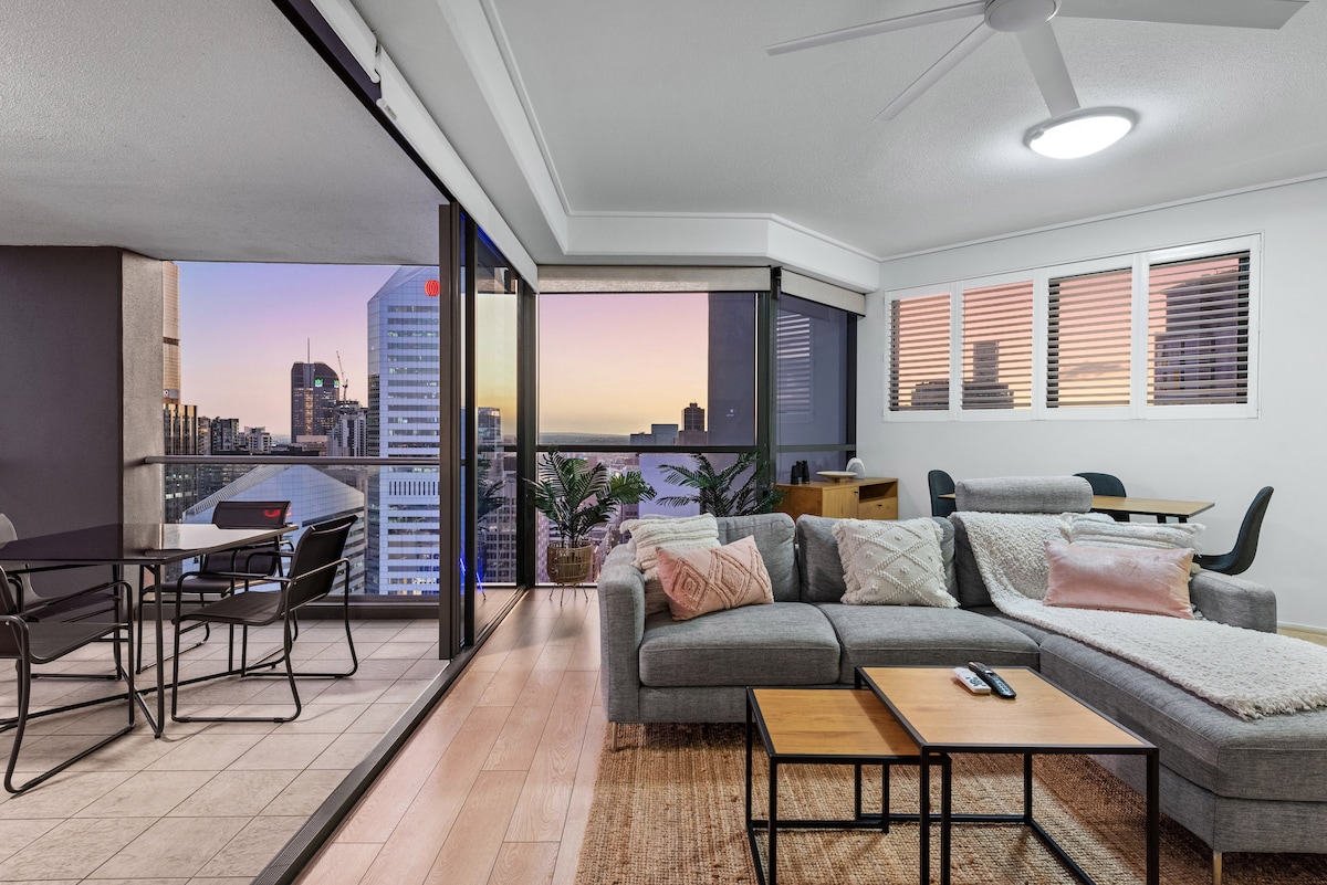 An open-plan living space features a modern, L-shaped sofa, a coffee table, and a dining area with chairs. Large glass doors provide access to the balcony, showcasing city views at dusk. Natural light fills the room through windows adorned with subtle blinds.