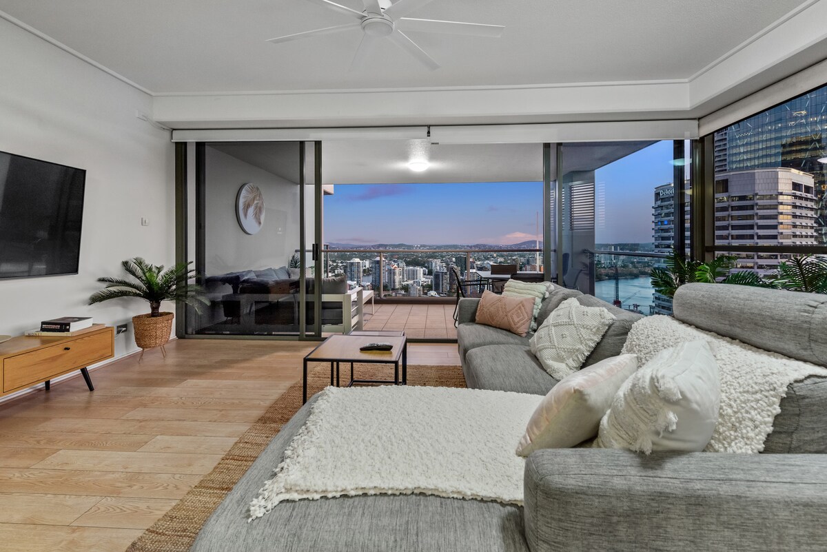A modern living area features a spacious gray couch with decorative pillows, a sleek media console, and a large wall-mounted TV. Floor-to-ceiling glass doors provide a view of the expansive balcony and cityscape beyond, creating a bright and inviting atmosphere.