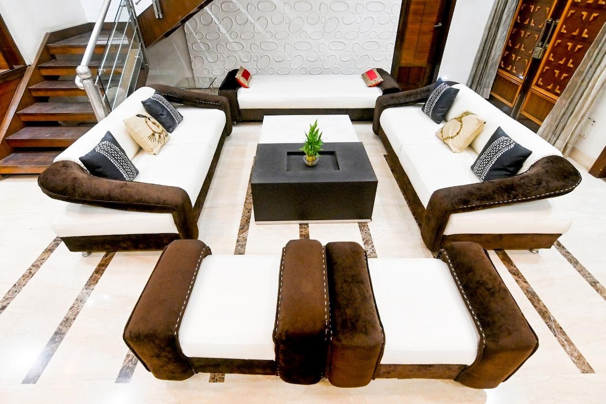A modern living area is arranged with two spacious sofas featuring white upholstery and dark brown accents. A minimalist coffee table holds a small potted plant in the center. Geometric designs on the wall add visual interest, while warm wooden elements provide contrast.
