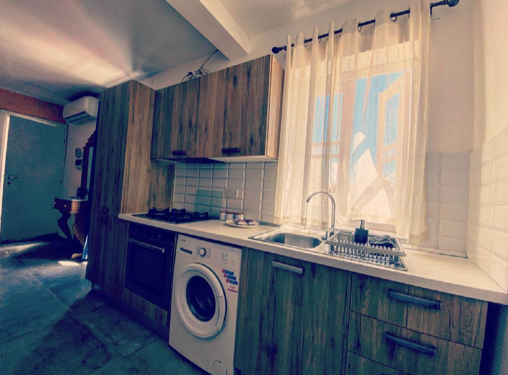 A compact kitchen features wooden cabinetry and modern appliances, including a washing machine and a gas stove. A window with sheer curtains allows natural light to brighten the space, reflecting off the light tile backsplash.