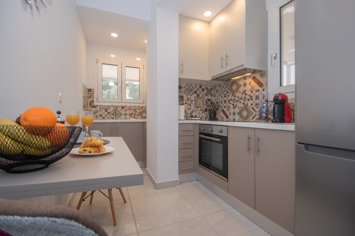 A modern kitchen is equipped with sleek cabinetry and an oven. A dining table is positioned nearby, set with a fruit bowl and pastries. Natural light enters through three windows, highlighting the patterned backsplash and stainless-steel appliances.