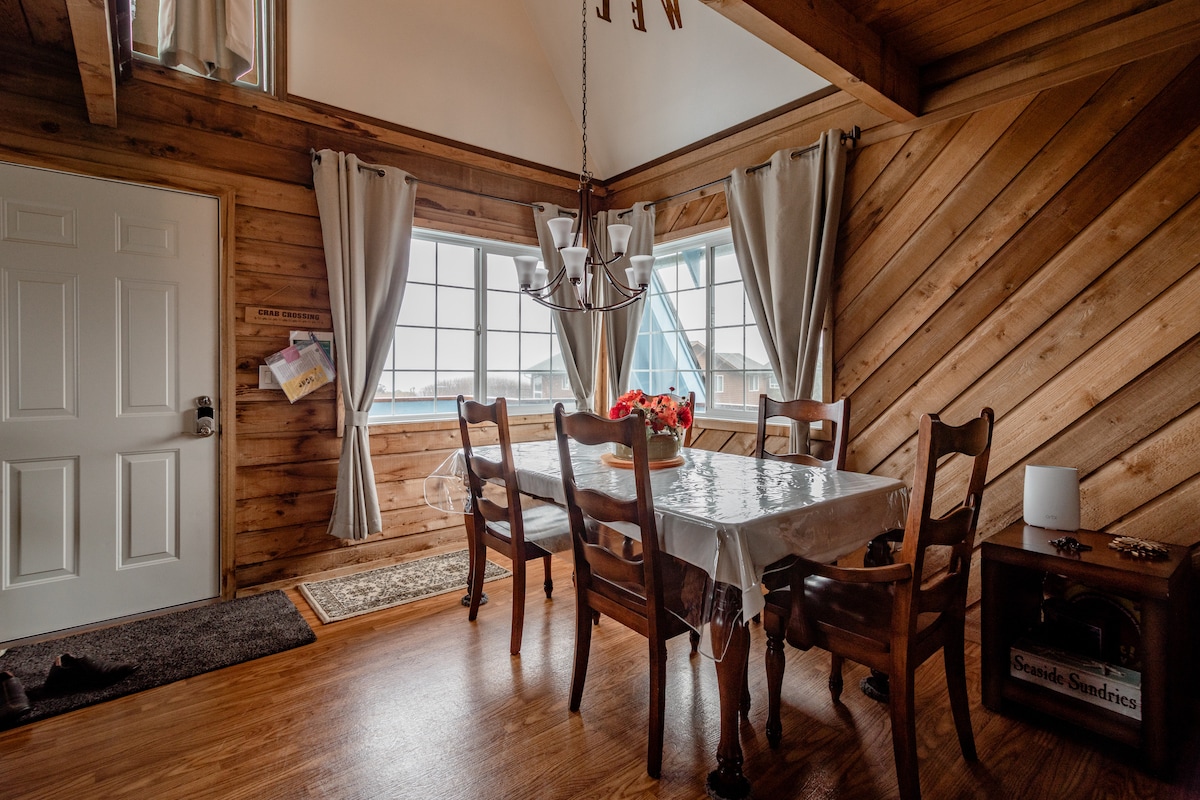 A cozy dining area features a large wooden table surrounded by six chairs. Natural light illuminates the space through two windows, adorned with light curtains. Cedar paneling creates a warm, inviting atmosphere, complemented by a chandelier hanging above the table.