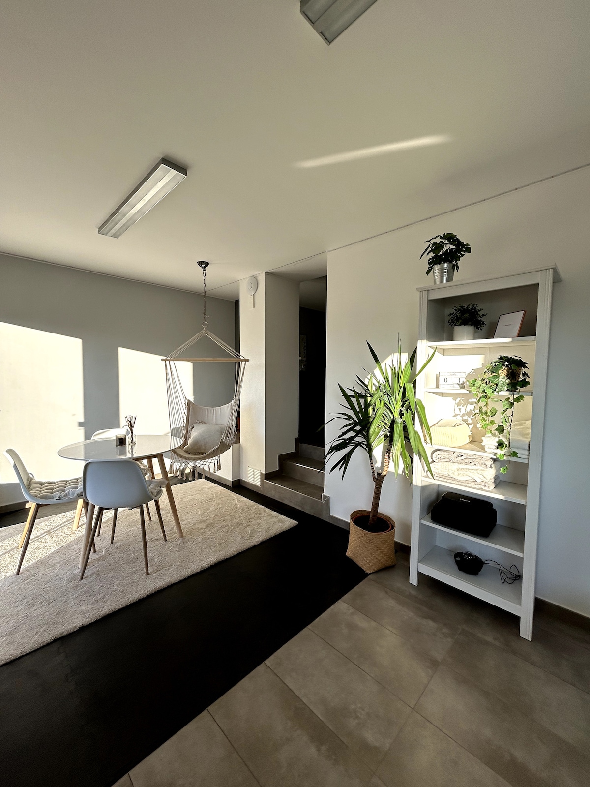 A well-lit corner of the living area features a circular dining table surrounded by white chairs. A hammock chair hangs nearby, while a tall plant adds greenery. A white bookshelf stands against the wall, showcasing decorative items and providing storage.