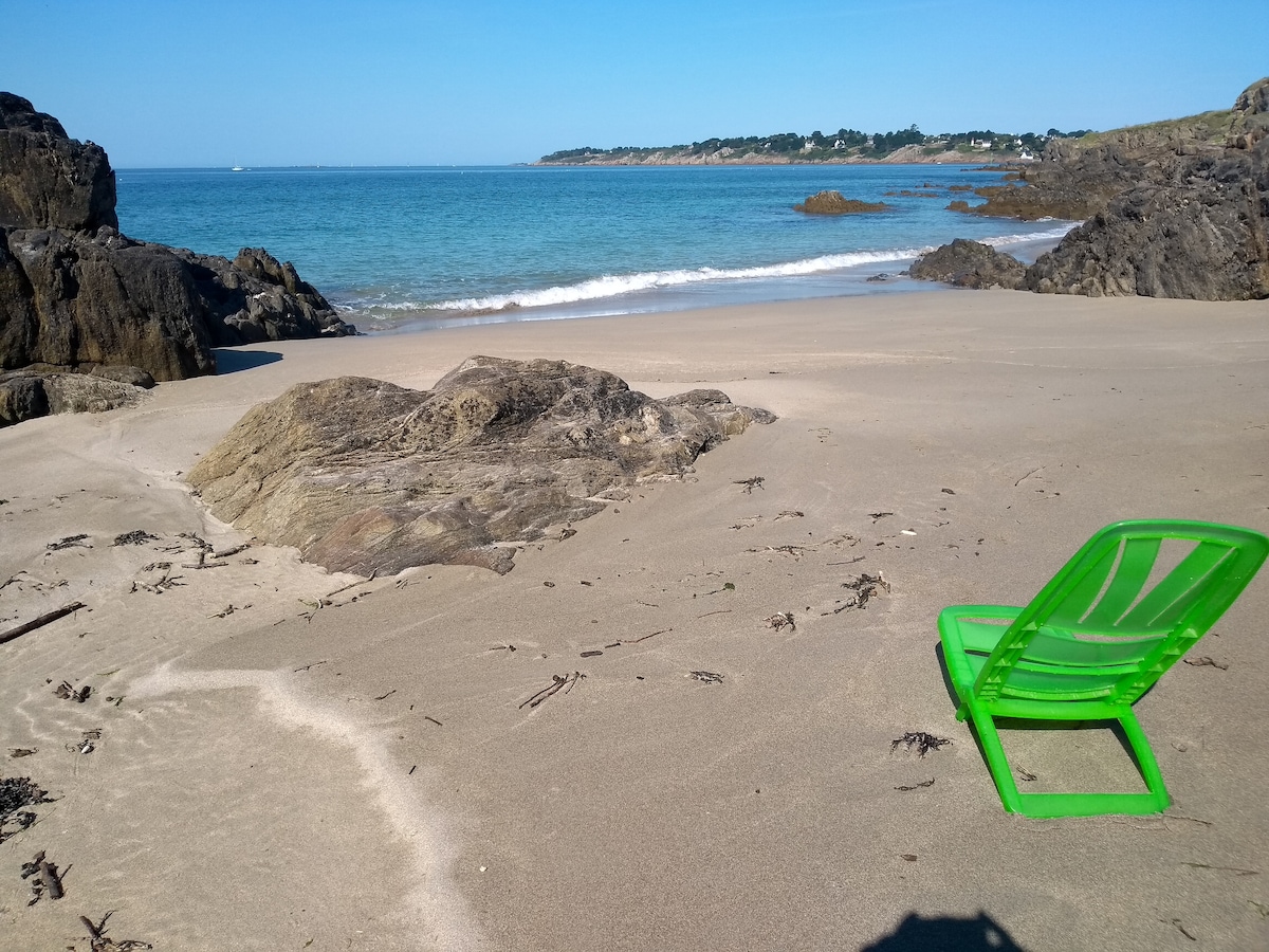 A tranquil beach scene is depicted, featuring a single green lounge chair placed on soft sand. Gentle waves lap at the shore, while a rocky coastline is visible in the background. Sunlight illuminates the calm water, creating a serene atmosphere.