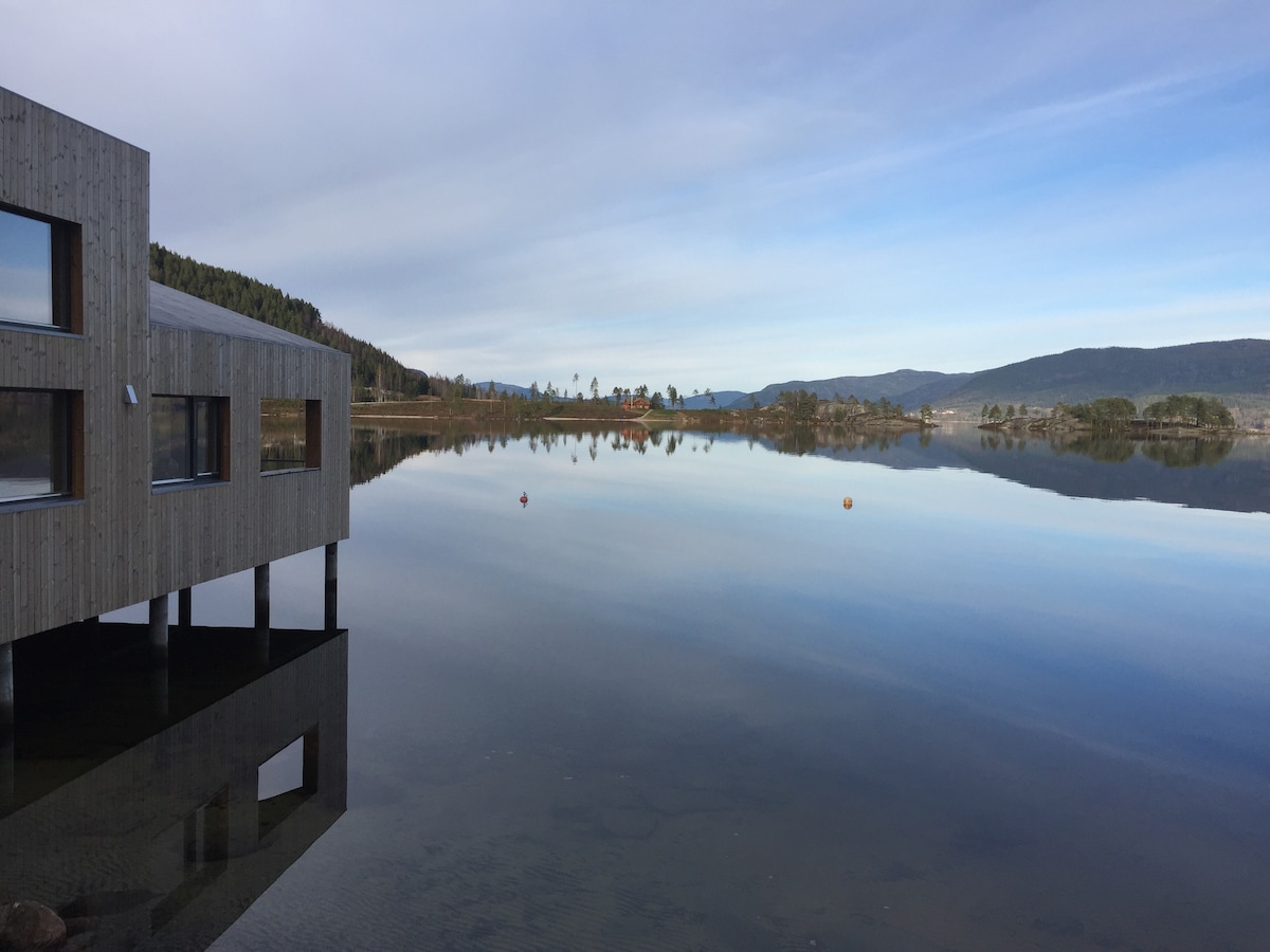A modern cabin extends over calm waters, with its wooden structure reflecting in the serene lake. The landscape features gently rolling hills and a clear sky, enhancing the tranquil setting, making it ideal for relaxation and connection with nature.
