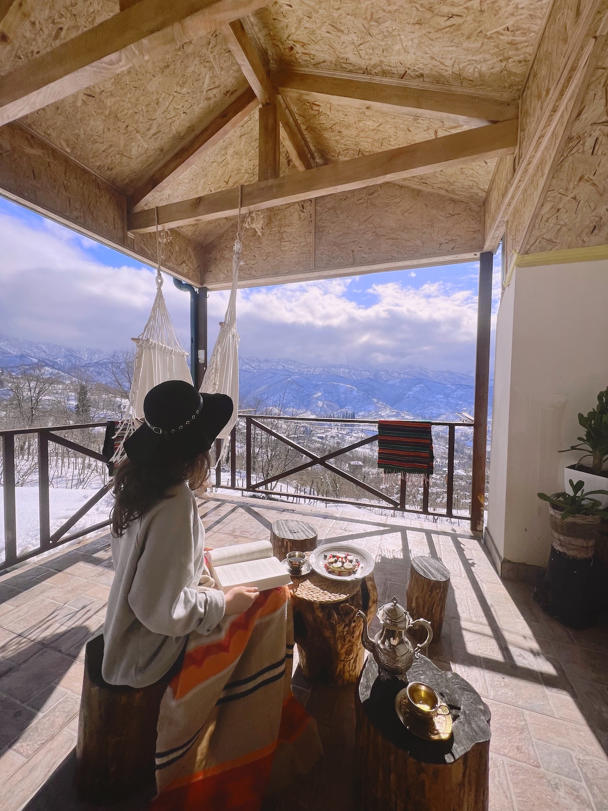 A cozy seating area features wooden logs as tables and stools, with a person reading a book. Hammocks gently sway in the background, and mountains are visible through large windows, surrounded by snow-covered landscapes.