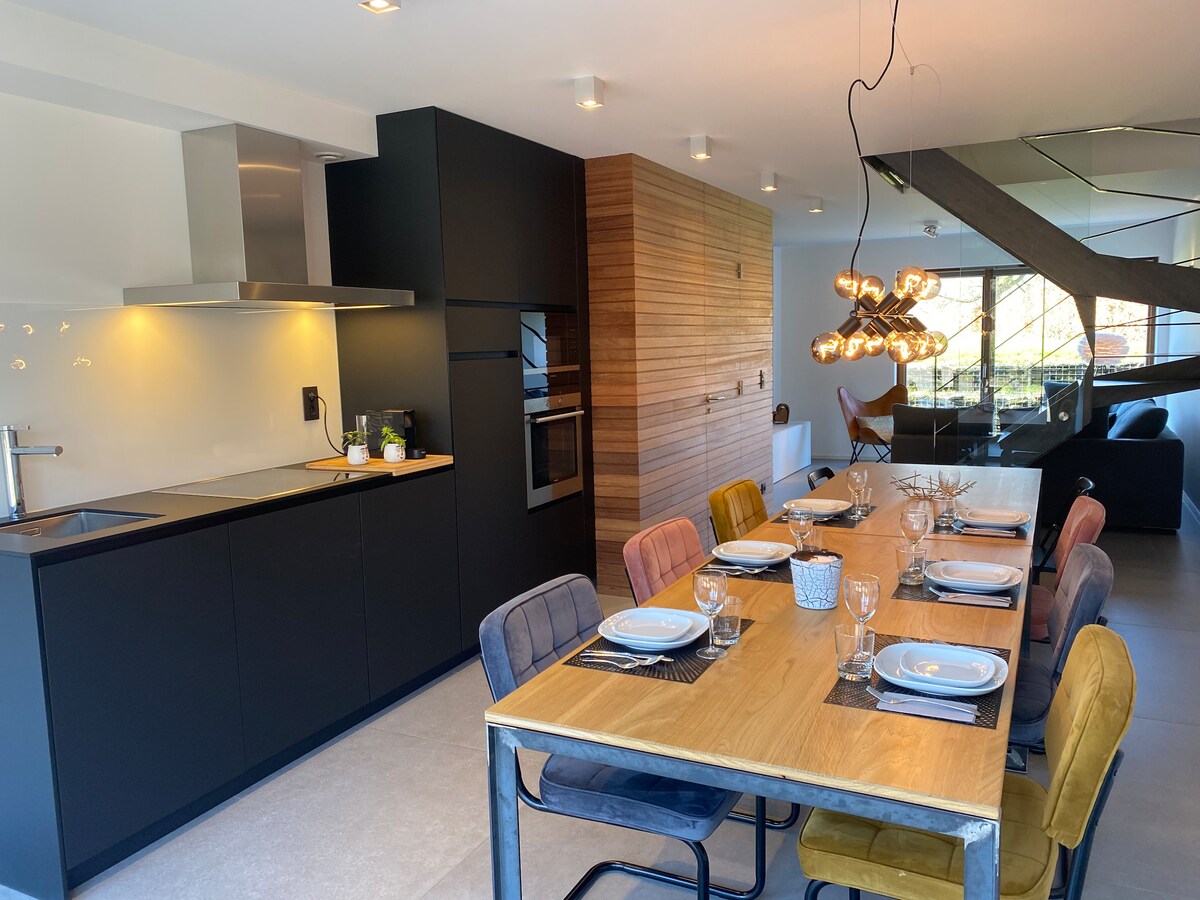 A modern kitchen area features sleek black cabinetry complemented by a wood-paneled wall. A dining table is arranged with plates and glasses, surrounded by chairs in various colors. Bright natural light streams in through large windows, enhancing the open and inviting space.