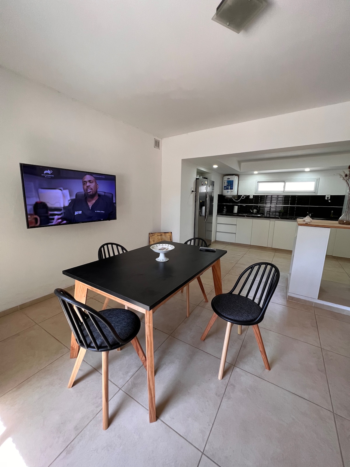 A modern dining area is featured with a black table surrounded by four wooden chairs. A bowl sits in the center of the table. A wall-mounted television is visible, and the open kitchen in the background includes sleek, white cabinetry.