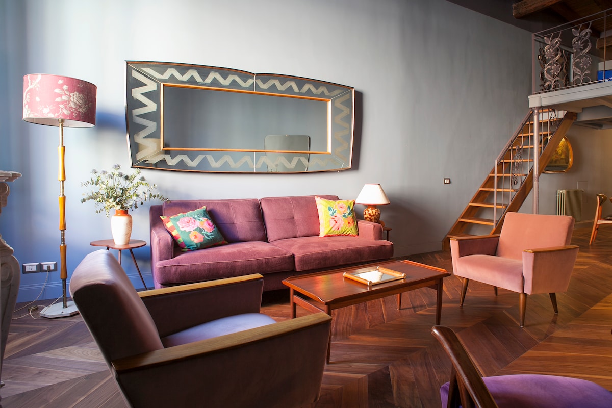 A spacious living area features a soft purple sofa adorned with vibrant throw pillows. Two comfortable armchairs provide additional seating, while a wooden coffee table sits centrally. A large mirror reflects the light, and a stylish lamp adds a touch of elegance to the room.