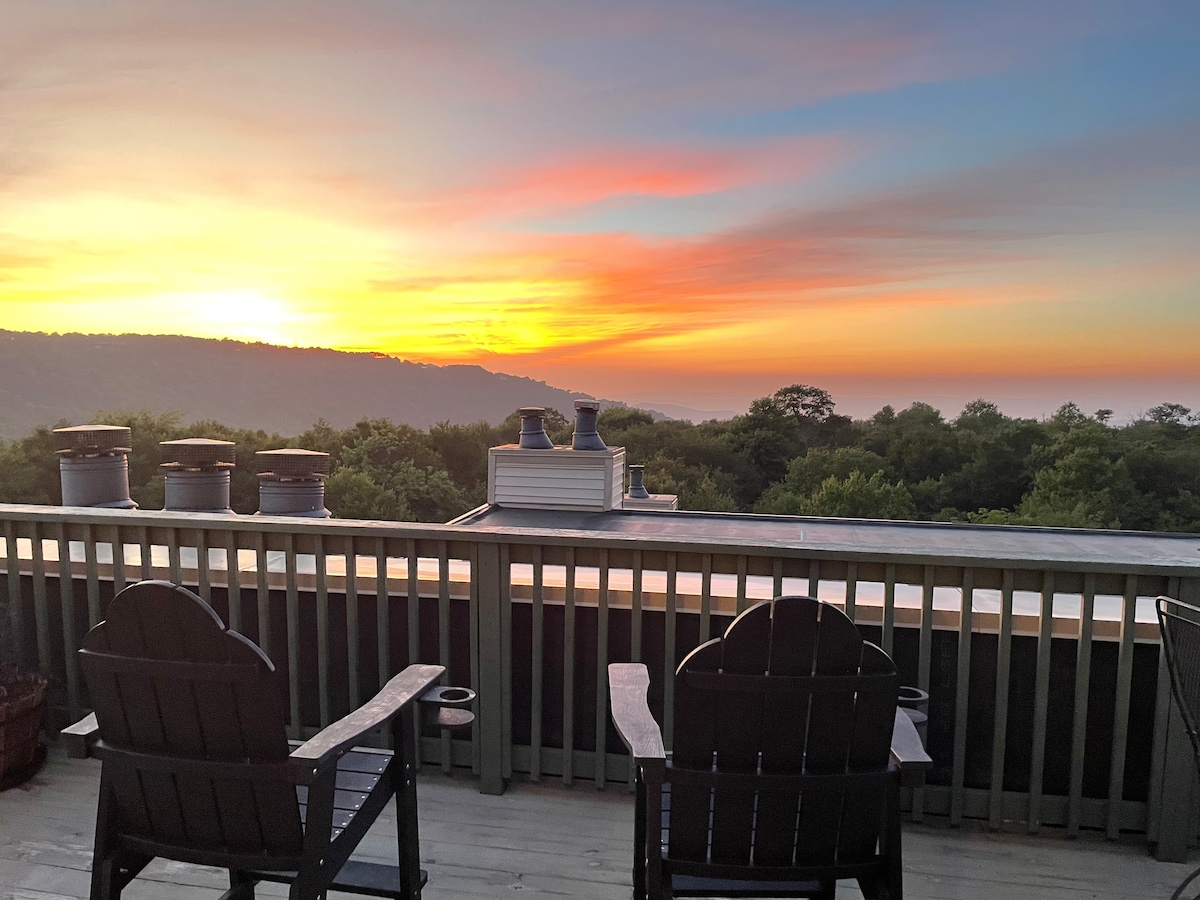 A tranquil view from the large private deck is presented, showcasing a vibrant sunrise over the mountains. Two black adirondack chairs are positioned on the deck, with greenery below and distant hills fading into the horizon.