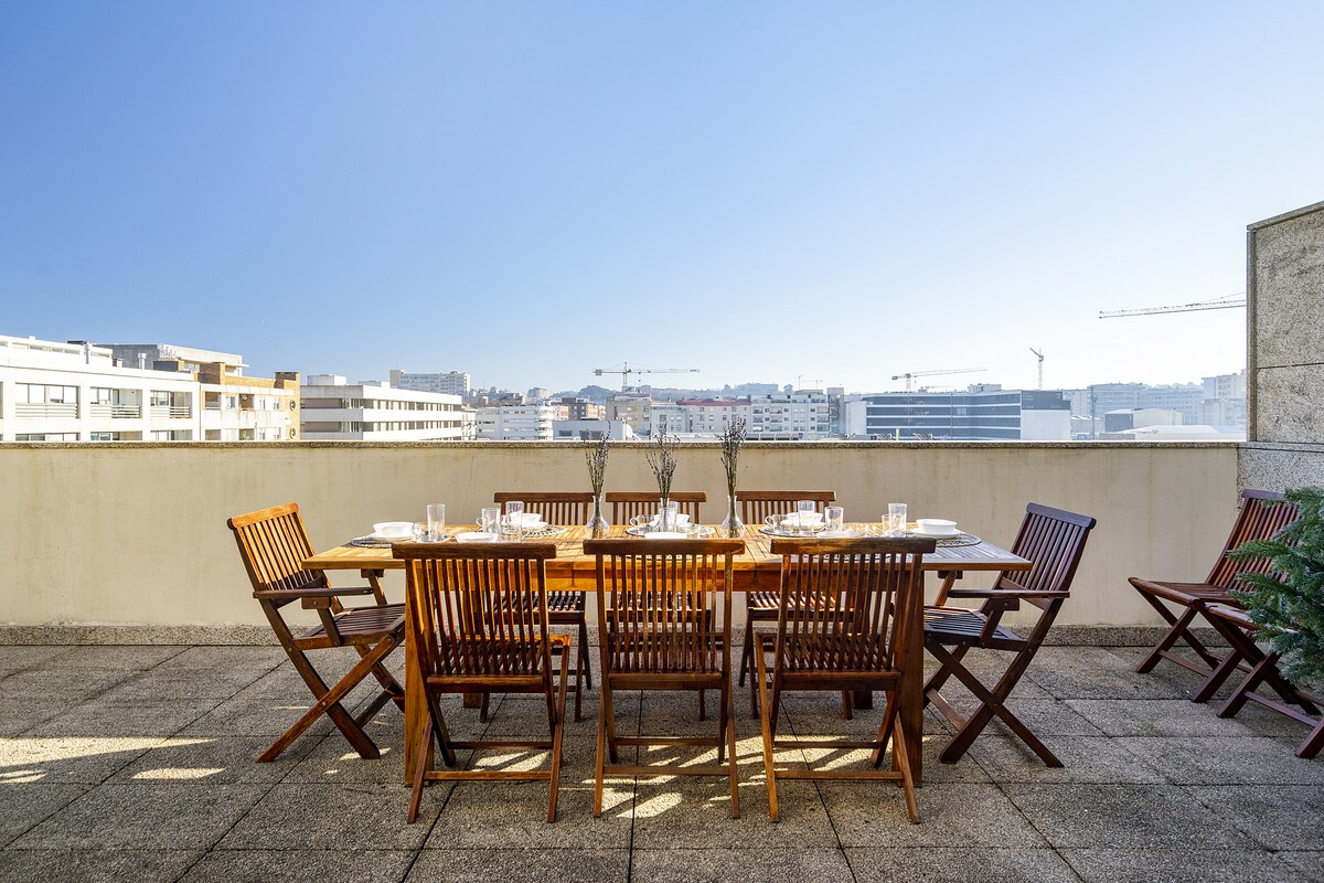 An outdoor terrace features a long wooden dining table surrounded by ten wooden chairs. The area is spacious with a tiled floor and an unobstructed view of the surrounding buildings and clear blue sky, ideal for family meals or gatherings.