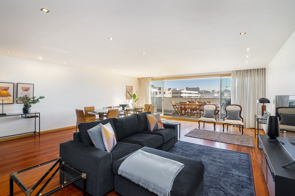A spacious living area features a large dark sectional sofa, complemented by a cozy throw. The room is illuminated by natural light from expansive glass doors leading to the terrace, showcasing a dining set and outdoor seating. Tasteful artwork adorns the wall beside a dining area.