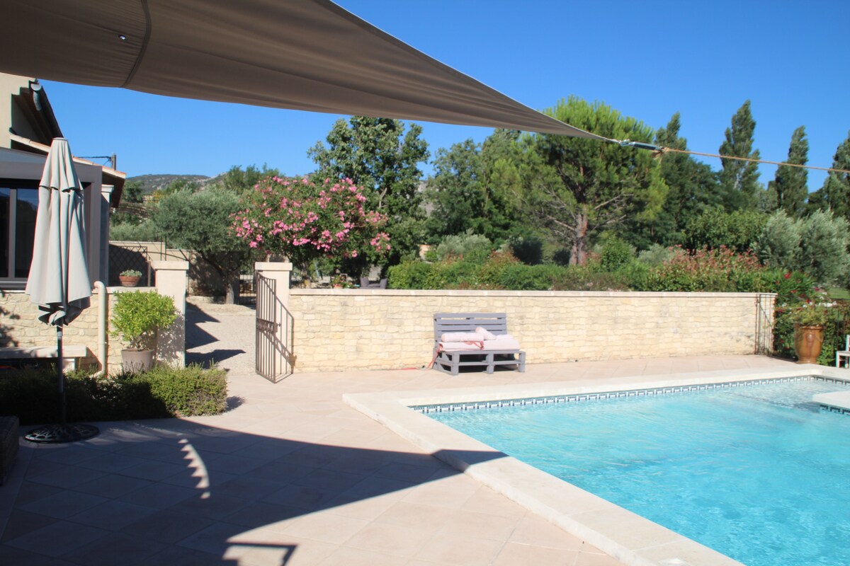 A private heated pool is surrounded by sun-drenched stone flooring, complemented by shaded areas provided by an awning. Lush greenery and flowering shrubs line the background, offering a serene garden view. A white lounge chair enhances the outdoor relaxation space beside the pool.