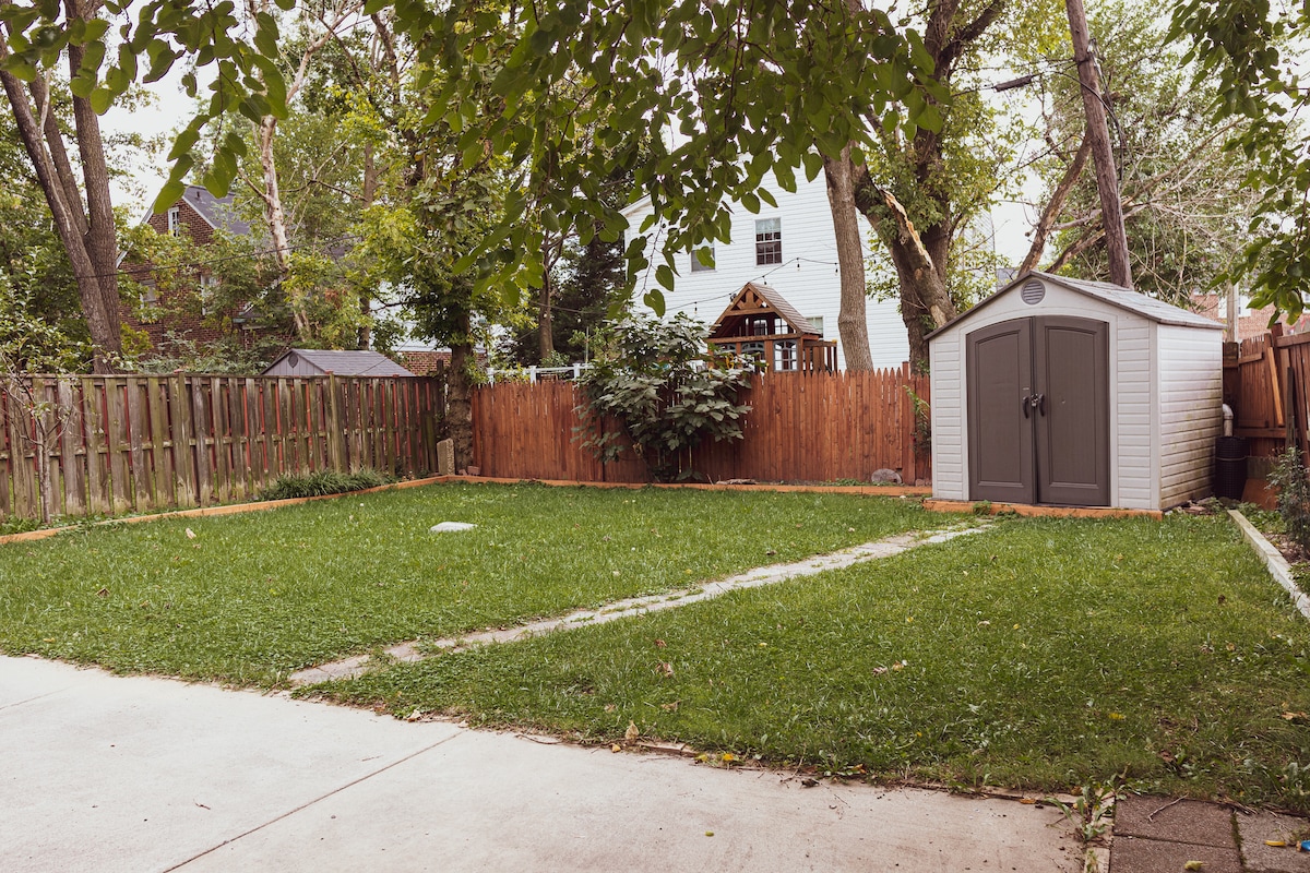 A spacious fenced backyard features a neatly maintained green lawn. A wooden playset stands in the background, surrounded by trees. A small storage shed is positioned to the right, complementing the serene outdoor environment.