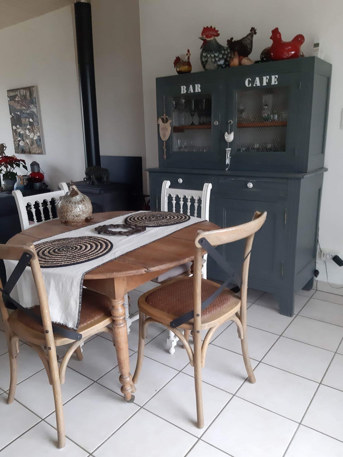 A wooden dining table is surrounded by two chairs, creating a cozy dining area. A dark grey cabinet serves as a storage piece, labeled 'BAR CAFE,' complementing the neutral tiled floor. Natural light highlights decorative elements, including table runners and a unique centerpiece.