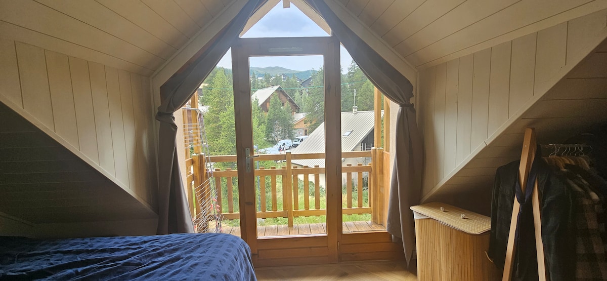 A spacious area showcases sloped ceilings and large glass doors leading to a wooden balcony. Natural light floods the room, illuminating a dark blue bedspread. Outdoor views of a forested landscape and neighboring structures are visible through the doors.