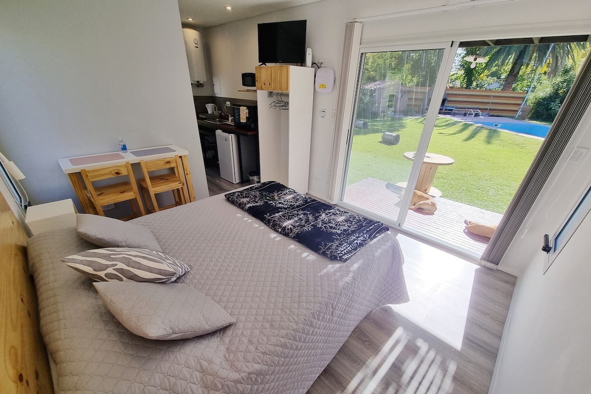 A bright sleeping area features a spacious bed adorned with a textured quilt and decorative pillows. Large glass doors allow natural light to fill the room and provide a view of the lush outdoor space with a pool and seating area.
