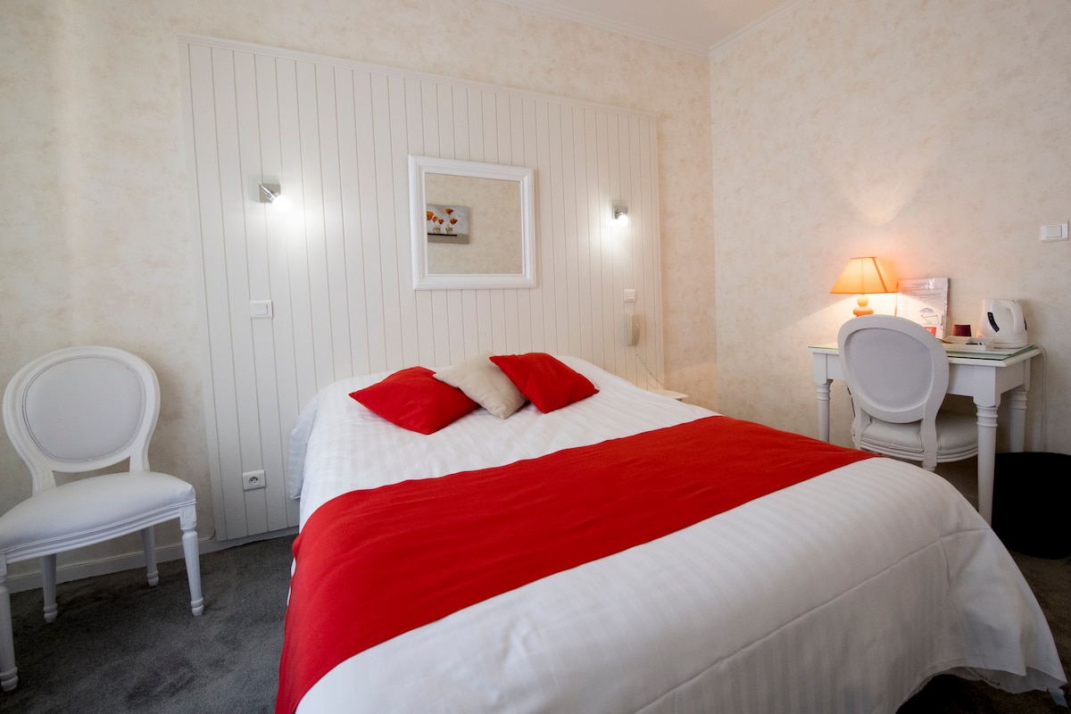 A bright and comfortable room features a spacious bed with a white and red coverlet. A small desk with a lamp is situated nearby, complemented by a mirror-mounted wall sconce. A simple chair is placed beside the bed, while soft carpeting completes the inviting ambiance.