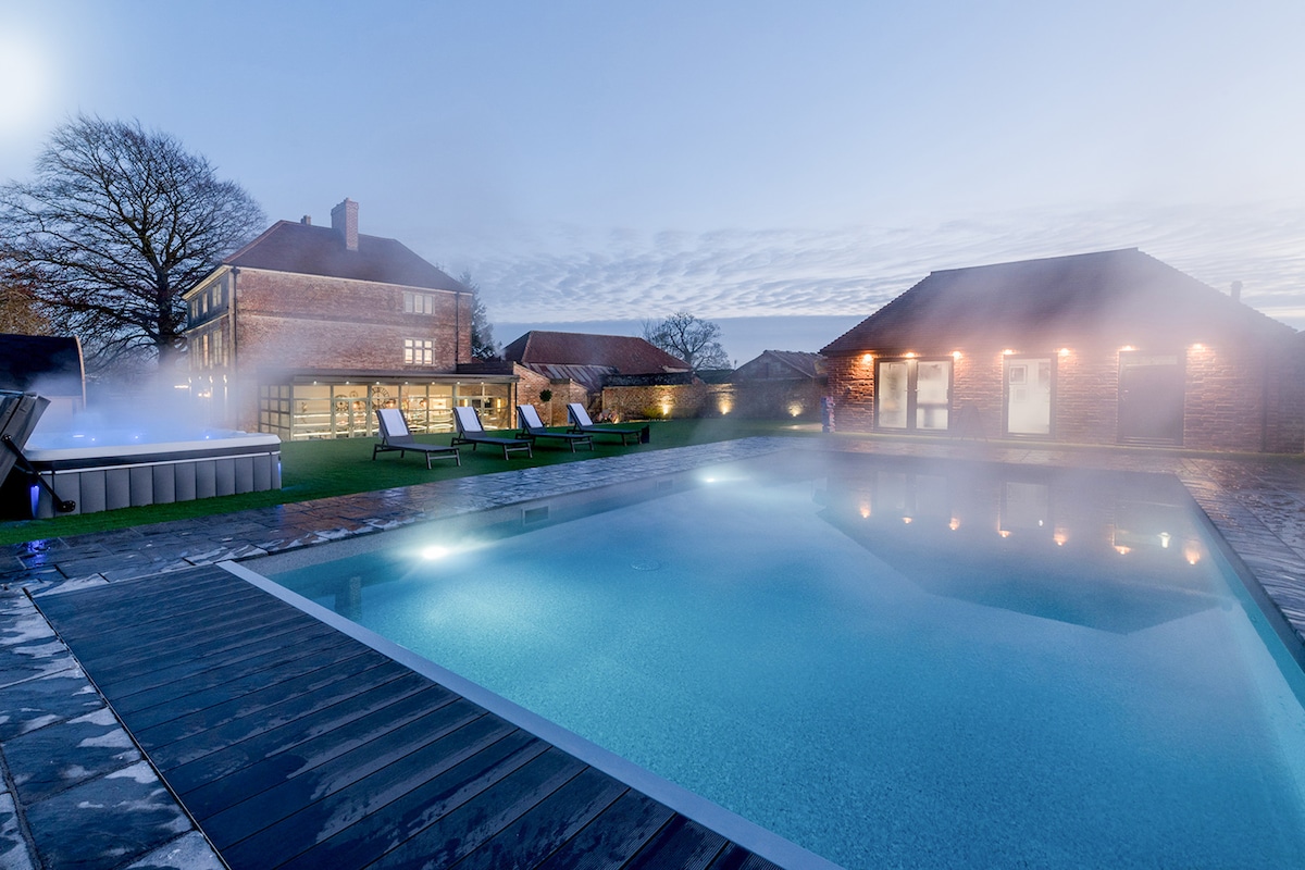 An outdoor heated pool is surrounded by a spacious sun deck, where sun loungers are arranged for relaxation. A hot tub is positioned nearby, with steam rising in the cool evening air. Soft lighting illuminates the pool area, enhancing the tranquil ambiance.