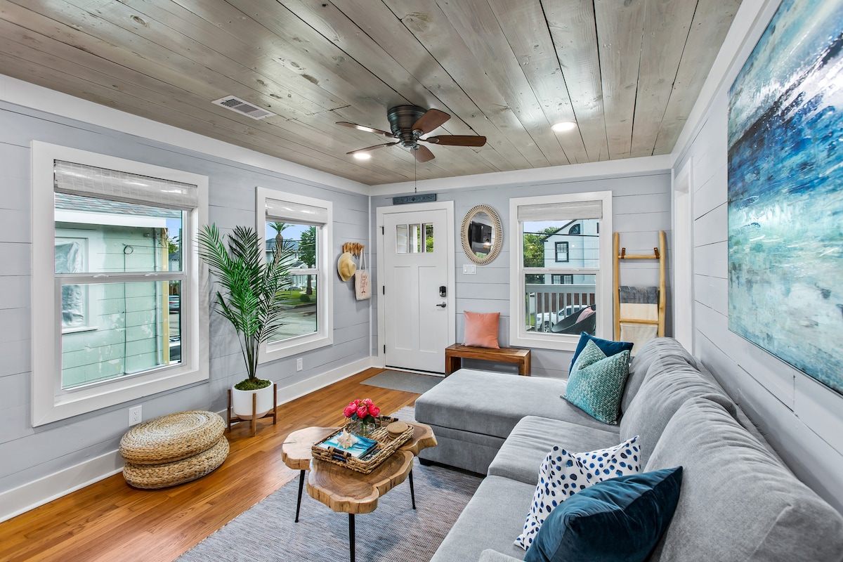 A welcoming living area features a spacious gray sectional sofa adorned with teal and patterned pillows. Natural light streams through multiple windows, illuminating the wooden flooring and shiplap ceiling. A wooden coffee table sits in front, holding a small floral arrangement and decorative items.