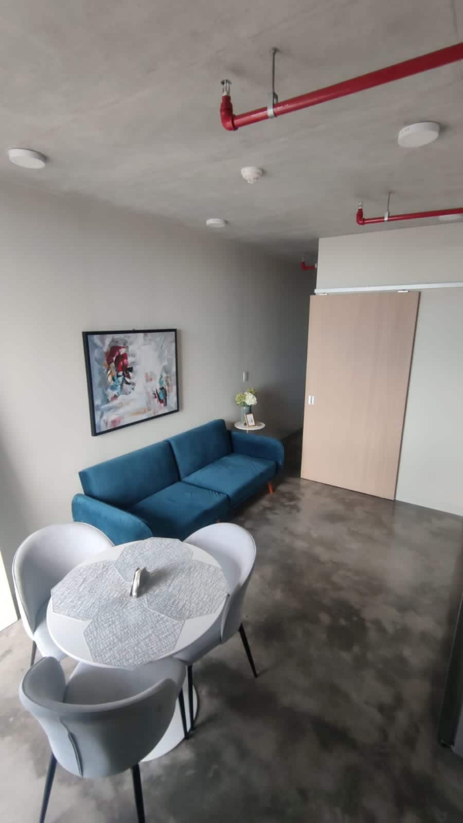 A spacious living area includes a blue couch positioned against a light gray wall, complemented by a modern artwork. A small round dining table is set for four, and a sliding door leads to another room. The polished concrete floor reflects the natural light.