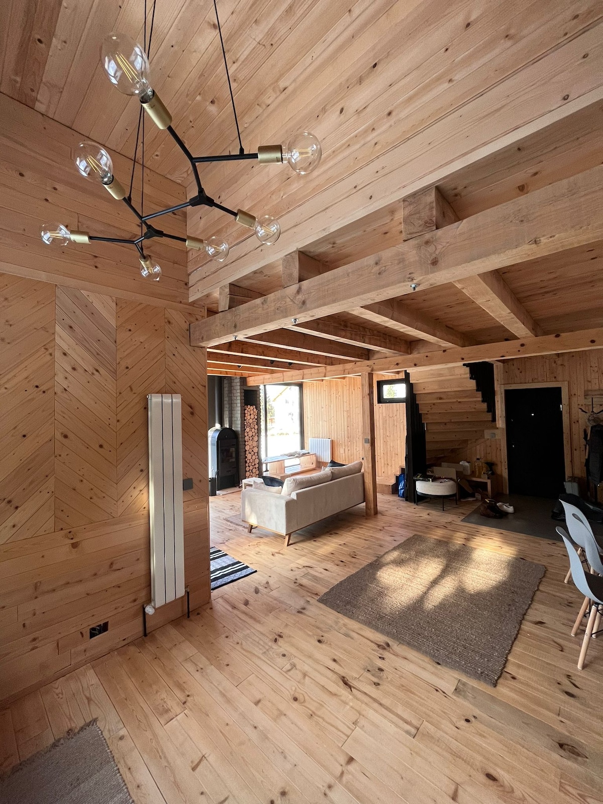 The interior features warm wooden walls and flooring, complemented by a contemporary chandelier. A cozy seating area is arranged near large glass doors, allowing natural light to fill the space. A staircase can be seen in the background, adding to the cabin's inviting character.