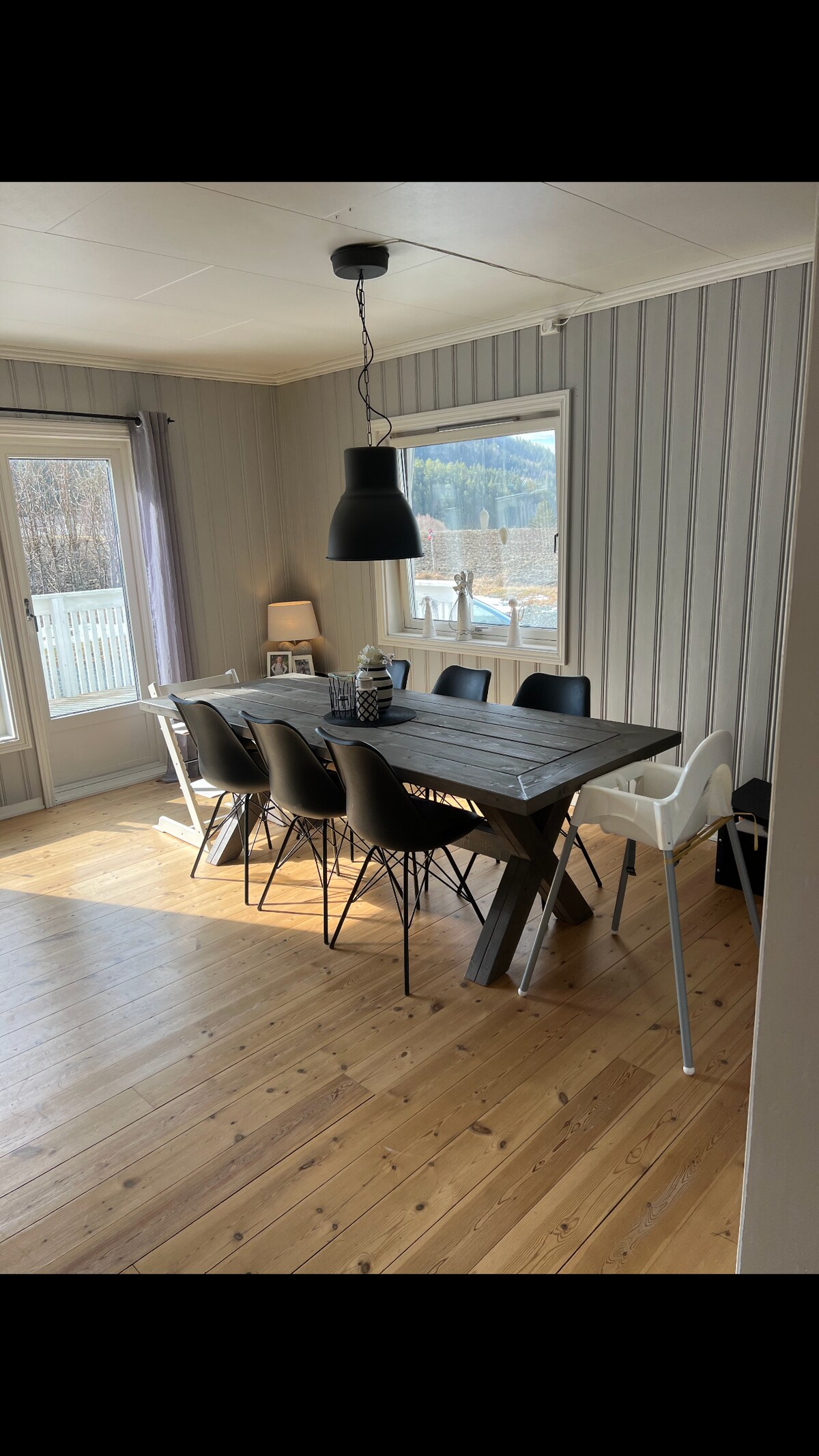 A dining area is presented with a large wooden table, surrounded by eight chairs in varying styles. A striking black pendant light hangs above, illuminating the space. Large windows offer views of the outdoors, while warm, wooden flooring enhances the room's inviting feel.