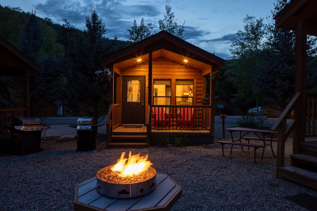 A rustic cabin is illuminated by warm exterior lights, with a welcoming porch and large windows showcasing the cozy interior. A fire pit with glowing flames is positioned in the foreground, surrounded by gravel. Lush trees and hills frame the background.