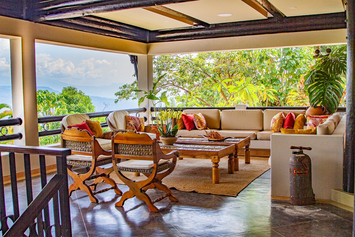 A spacious seating area features a large corner sofa adorned with colorful cushions, complemented by two wooden armchairs. A central wooden coffee table is surrounded by a natural fiber rug. Large windows provide views of lush greenery and distant mountains.