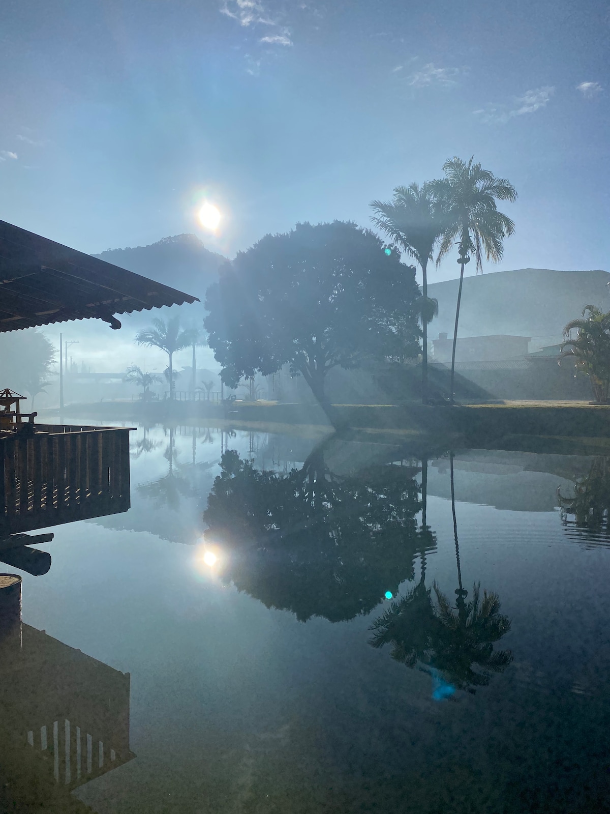 A calm pond reflects the sun and surrounding trees, creating a serene atmosphere. Mist gently envelops the area, while a covered structure is visible beside the water, enhancing the tranquil setting.