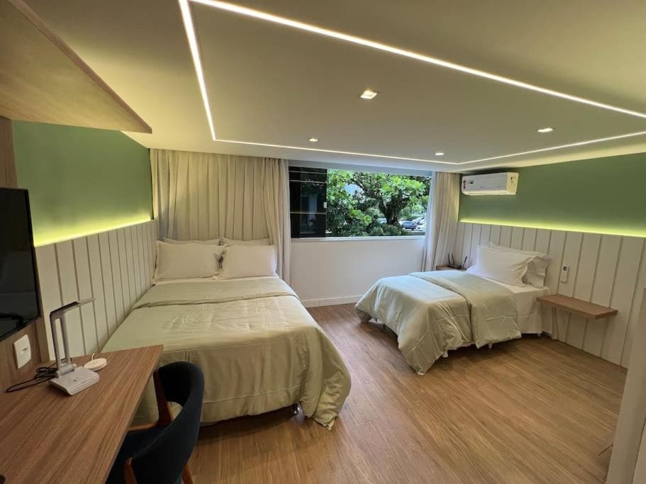 The suite features two beds: a spacious double and a single, both dressed in light-colored linens. A modern desk and chair are positioned against one wall, while large windows allow natural light to fill the room. Air conditioning units are mounted for added comfort.