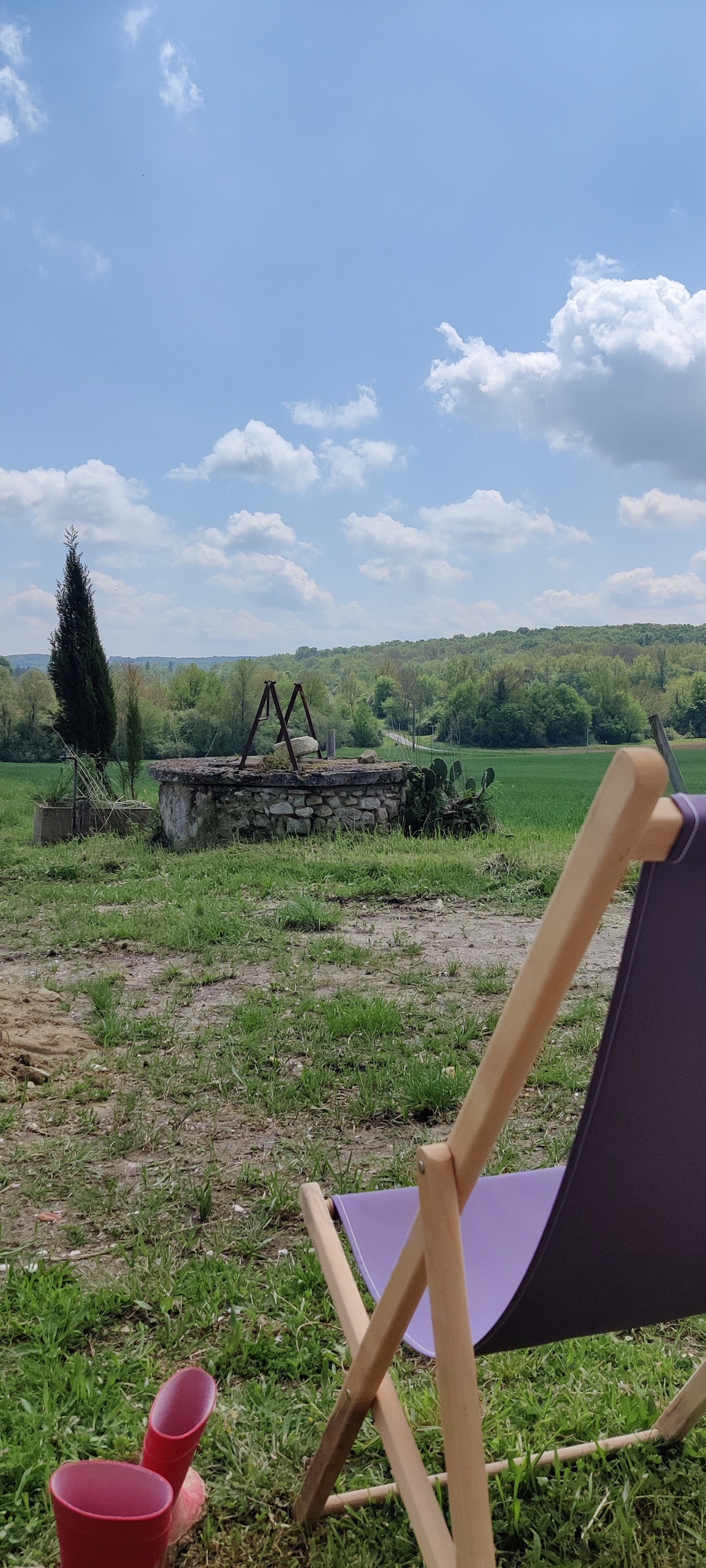 A serene outdoor scene is presented, featuring a wooden chair facing a vast green landscape. A stone well is seen in the background, surrounded by lush greenery and trees under a bright blue sky with scattered clouds.
