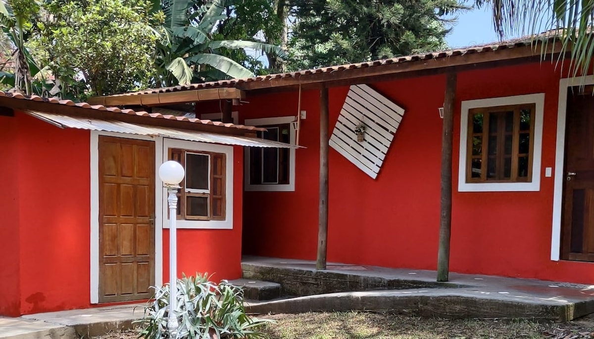 A vibrant red exterior is complemented by wooden doors and windows, framed by lush greenery. A small structure is visible to the left, with a white lamp post situated nearby, adding to the inviting appearance of the property.