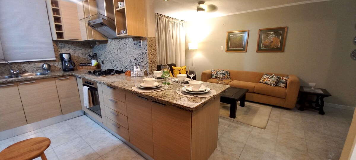 A modern kitchen is featured with stylish wooden cabinetry and granite countertops. An island counter is set for dining, complemented by a comfortable living space with a sofa and decorative cushions. Natural light brightens the area through large windows, enhancing the inviting atmosphere.