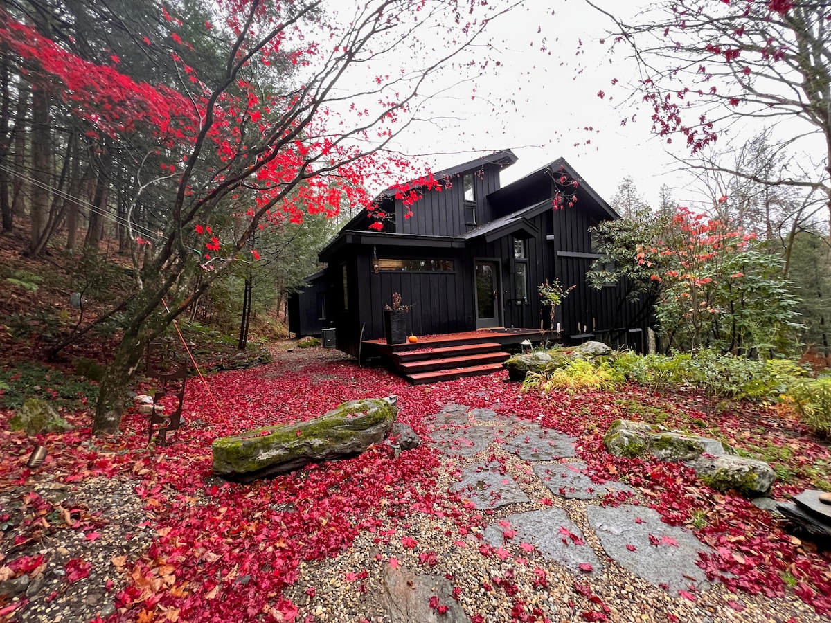 The exterior of the home is surrounded by vibrant red foliage, with scattered leaves covering a stone path leading to a deck area. Dark wooden siding contrasts with the natural landscape, and the house is nestled among trees, enhancing a sense of seclusion.