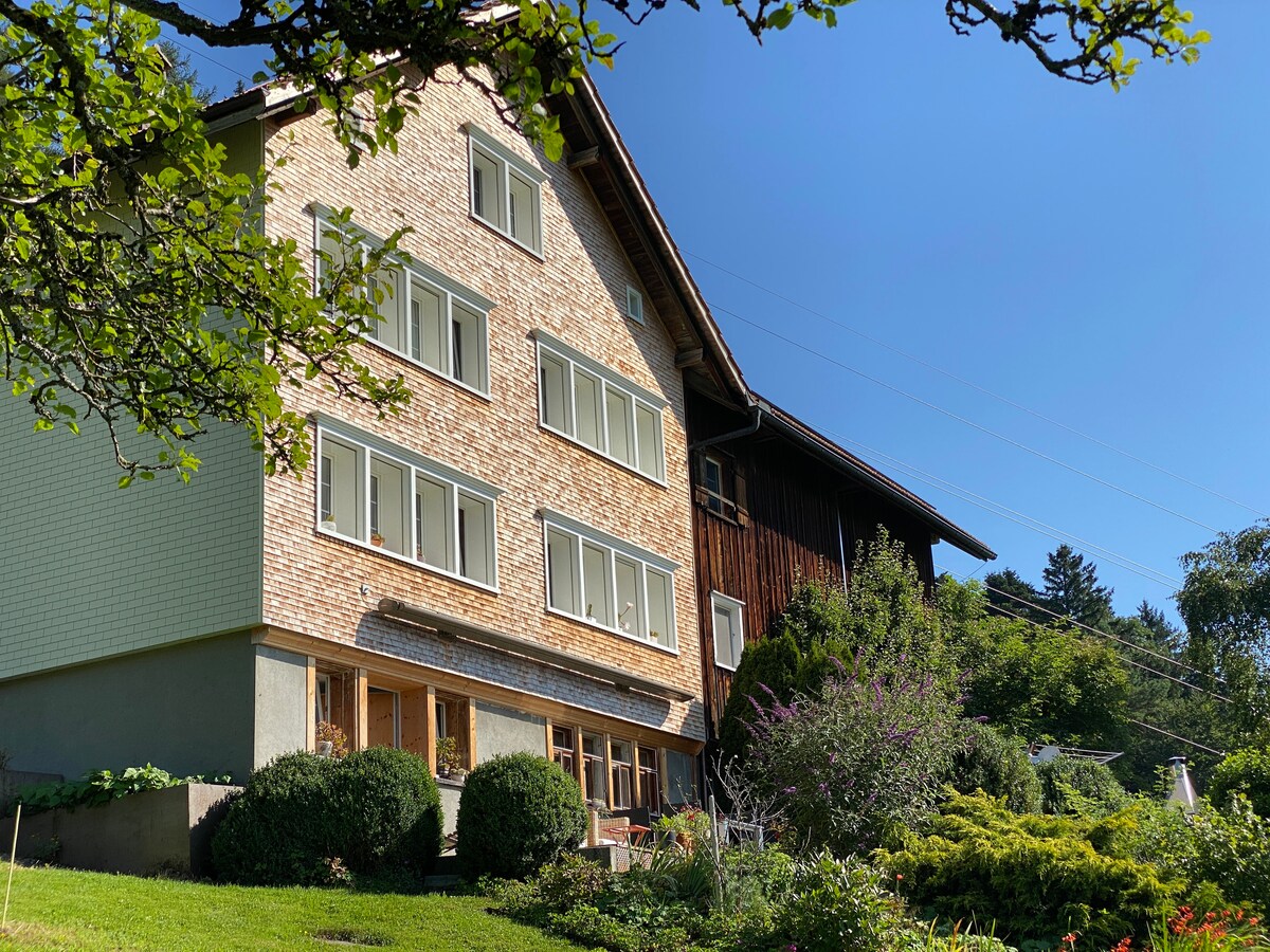 The image features a charming three-story Appenzeller house with a light-colored brick facade and large windows. A wooden structure is shown alongside, surrounded by lush greenery and garden areas. The clear blue sky enhances the inviting atmosphere of the rural setting.