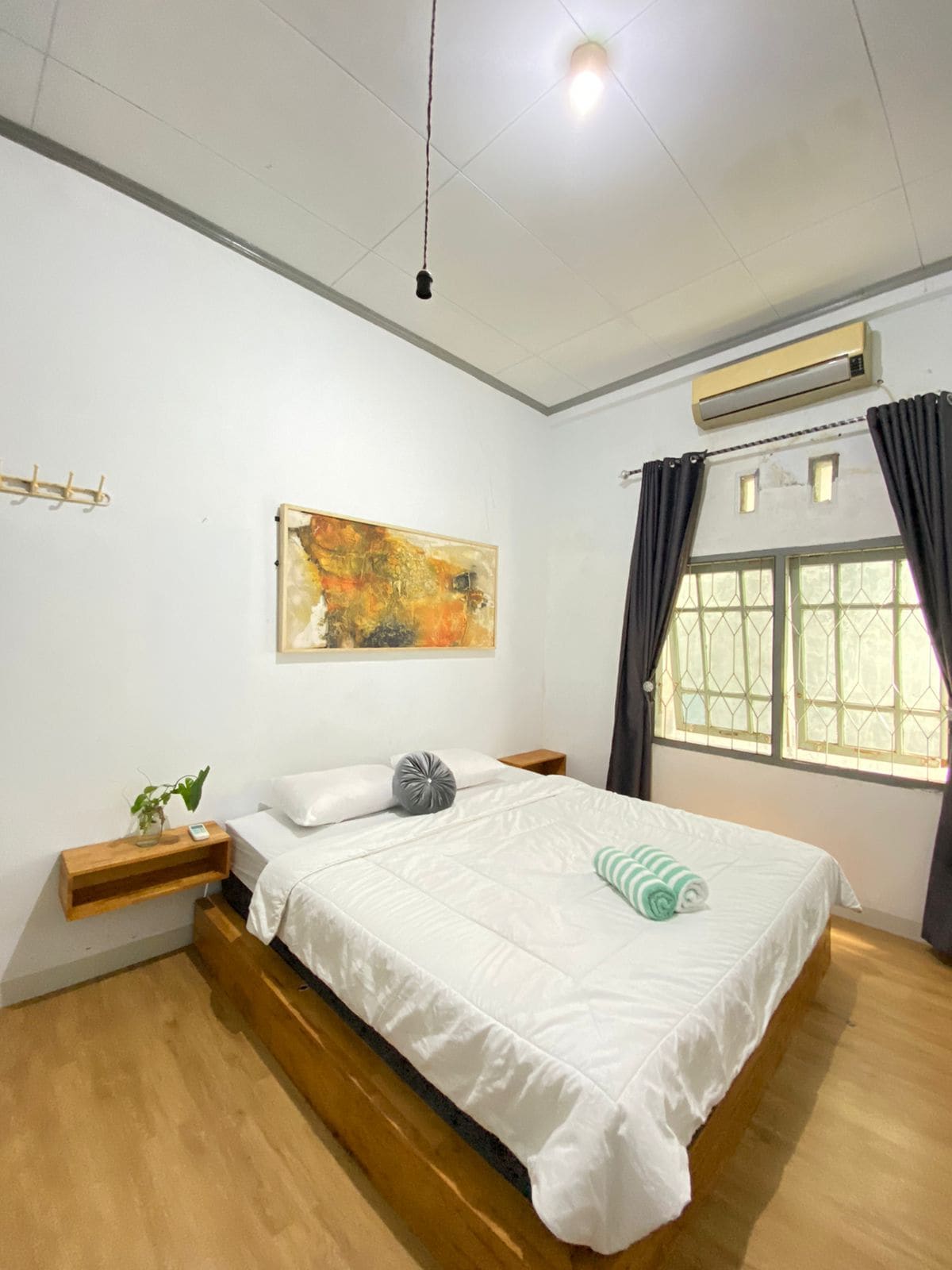 The room features a spacious bed with white bedding, complemented by a decorative pillow and a rolled towel. A piece of abstract art adorns the wall, and natural light filters through the window, framed by dark curtains. Air conditioning is also available.