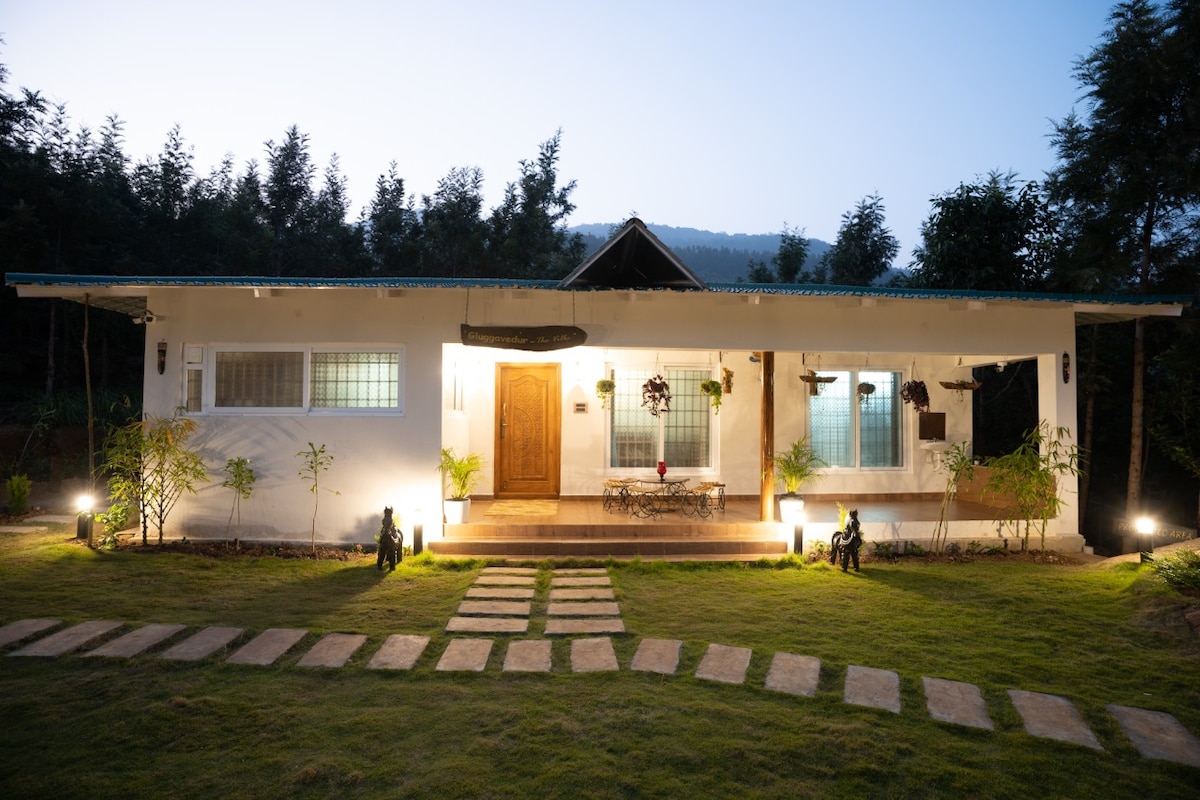 A single-story villa is framed by a well-maintained garden. Clear pathways lead to the entrance, illuminated by soft lighting. The facade features large windows on either side of a central wooden door, providing a bright and welcoming appearance against the backdrop of a mountain landscape.