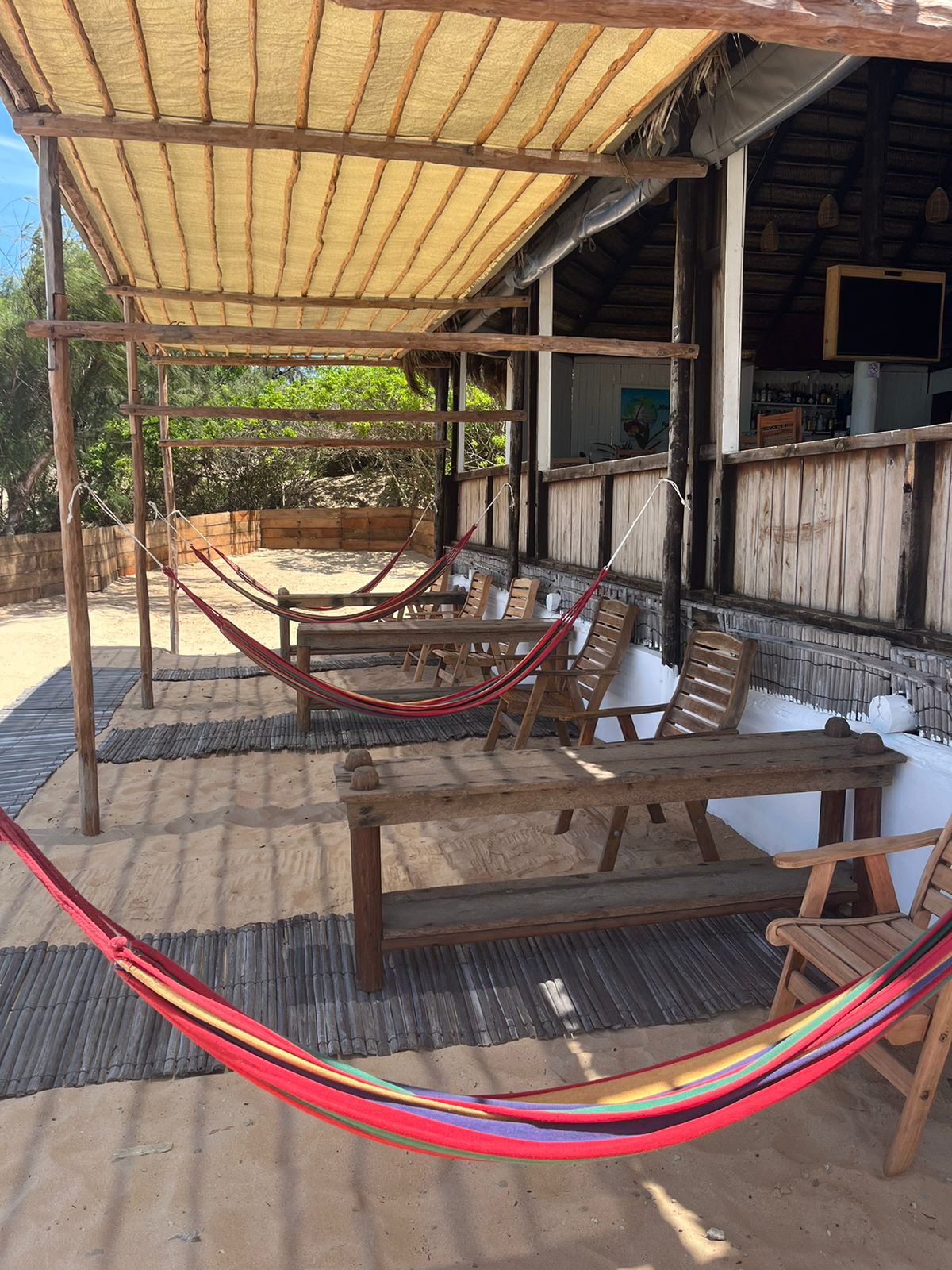 A shaded area features multiple hammocks suspended between wooden posts, alongside several wooden tables and chairs. The sandy ground complements the relaxed atmosphere, creating a space for leisure and socialization. Natural light filters through the overhead canopy.