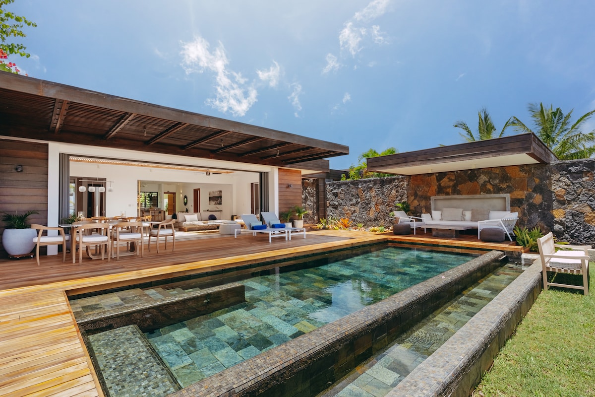 A modern villa showcases a wooden terrace extending towards a crystal-clear pool. Spacious seating areas are visible, with sunlight illuminating the bright outdoor space. Surrounding tropical greenery enhances the environment, while a cozy lounge is positioned near the edge of the pool, inviting relaxation.
