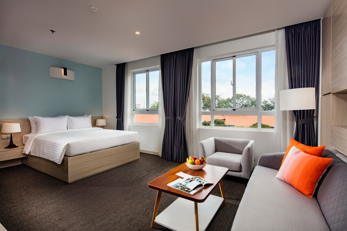 A spacious room is showcased, featuring a queen bed with crisp linens. Two large windows provide natural light and views of the outdoors. A seating area includes a gray armchair and a sofa with orange accents, complemented by a coffee table displaying fruit and magazines.