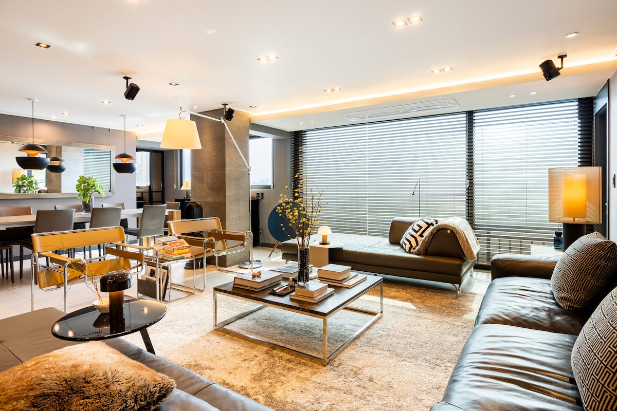The spacious living room features modern furniture with a combination of leather seating and a decorative coffee table. Large windows allow natural light to fill the room, while a neutral color palette is complemented by stylish decor. An inviting dining area is visible in the background.