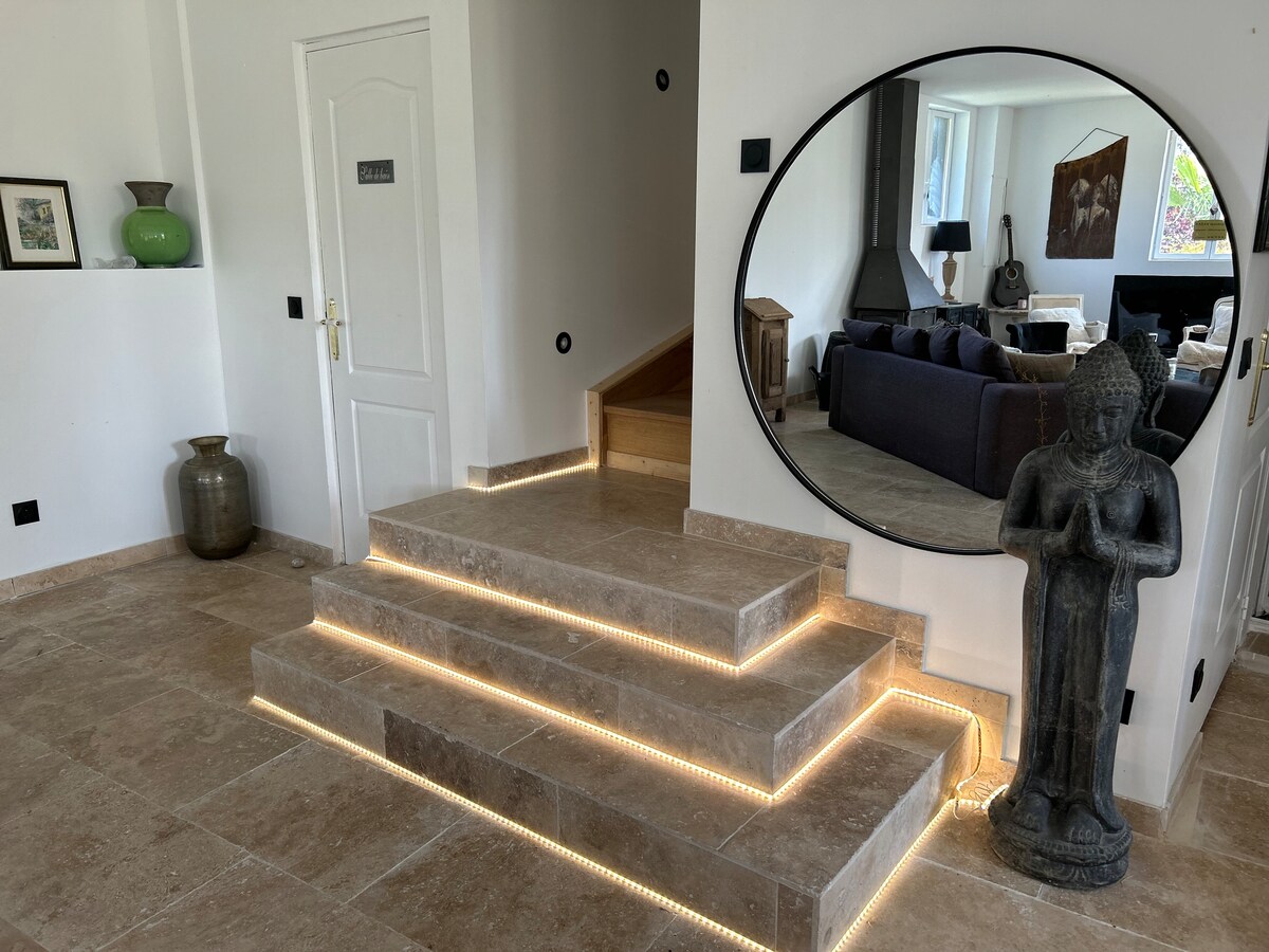A well-lit entryway features stone steps leading to an upper level. A round wall mirror reflects the living area, which displays a dark sofa. A decorative statue stands beside a potted plant, adding character to the space.