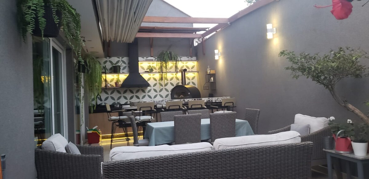 An outdoor dining area is presented with a long table surrounded by dark wicker chairs. Greenery adorns the walls, complementing the modern kitchen space with a decorative backsplash. Soft lighting creates a welcoming ambiance in this spacious area, ideal for family gatherings.