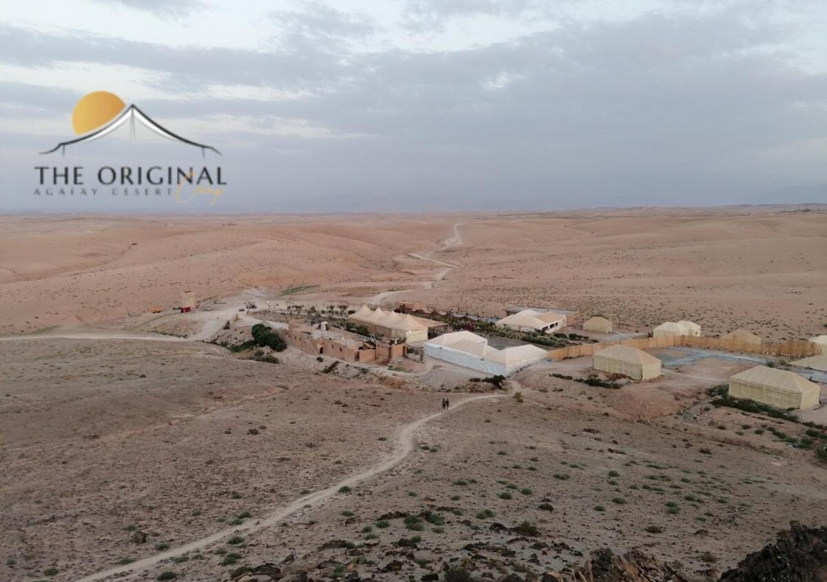 A panoramic view showcases the expansive rocky landscape of the Agafay desert. Tents are positioned along a dusty path, blending with the earthy tones of the surroundings. Gentle undulations of sand dunes stretch across the horizon under a soft, overcast sky.