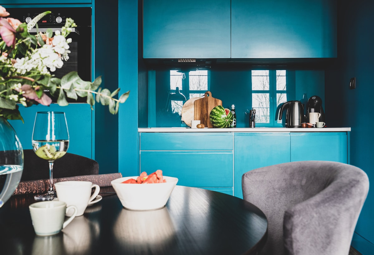 A modern kitchen area showcases vibrant blue cabinetry and a dark round table set with fresh fruit and glassware. A kettle and kitchen utensils are visible, along with a cutting board. Light streams through the windows, enhancing the lively atmosphere.