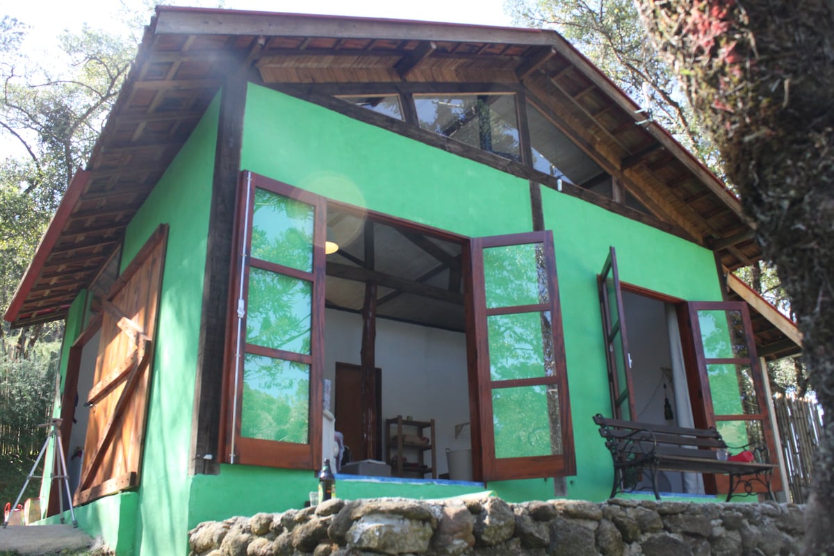 The exterior of a rustic green chalet is shown, featuring large open windows that invite natural light. A wooden bench in front offers a view of the surrounding nature, while stone accents complement the building's charm.