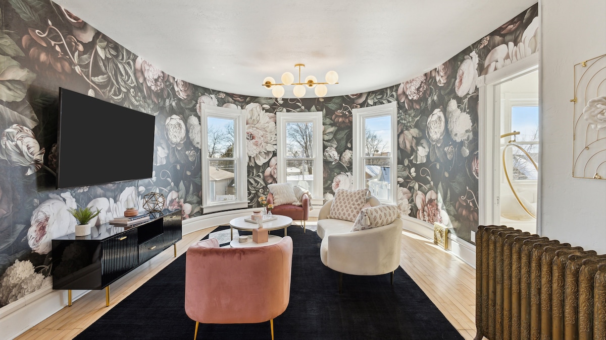 A stylish living area features a curved wall adorned with floral wallpaper. Two plush armchairs in light tones flank a circular coffee table, while a sleek black entertainment unit houses a flat-screen television. Large windows provide natural light, complementing the warm wooden flooring.
